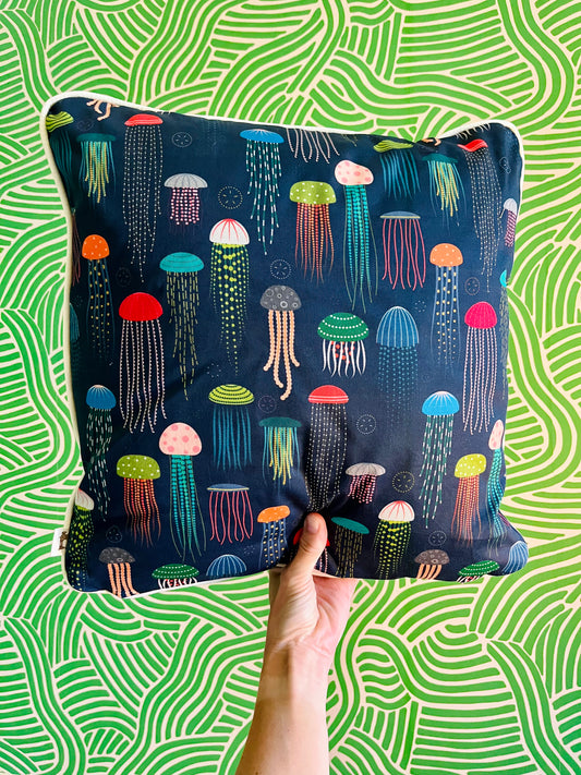 Photo of cushion in a dark blue fabric covered with a multicoloured jellyfish pattern. Cushion held in front of a wiggly green background